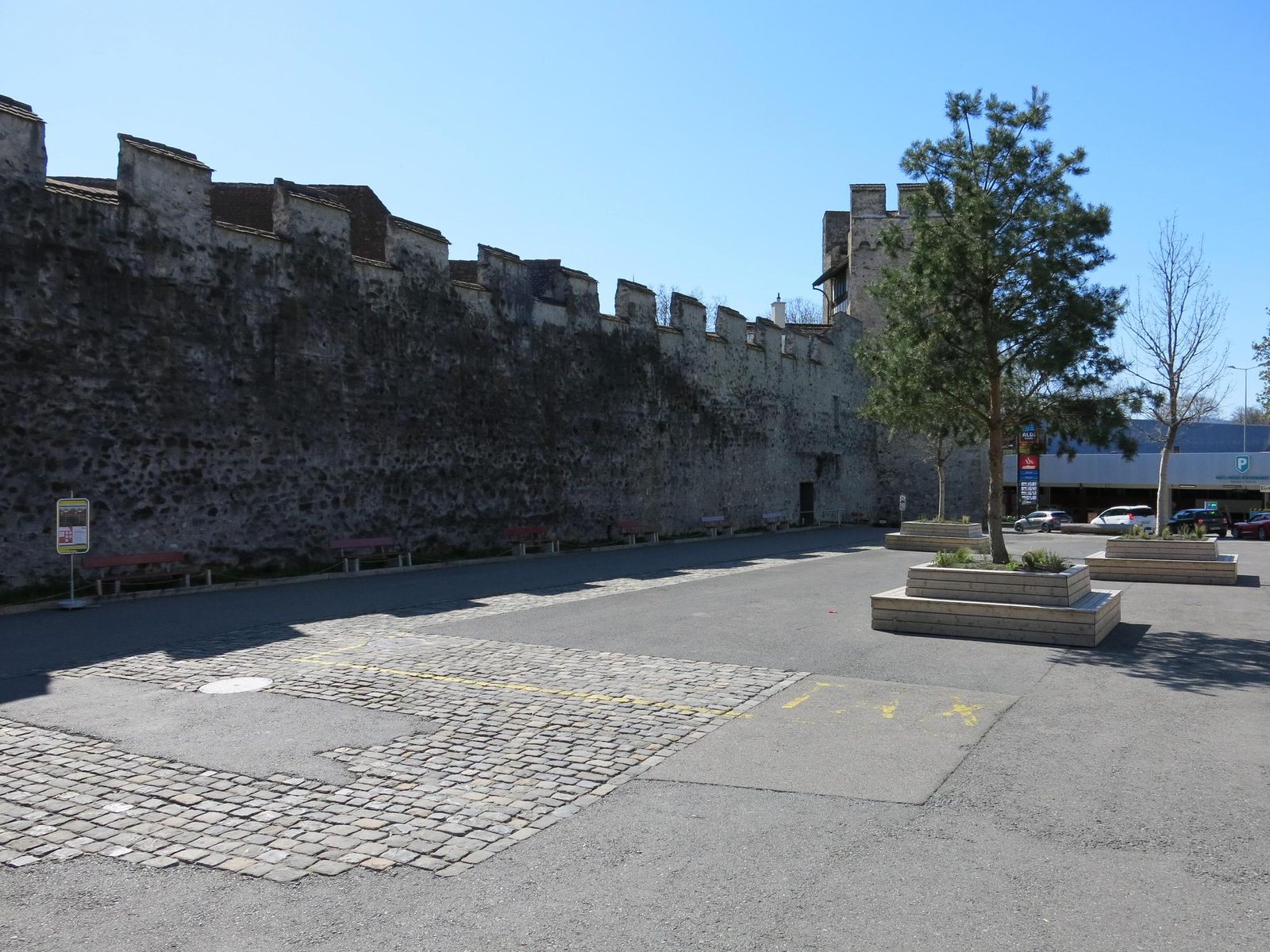 Der Platz an der Stadtmauer mit Turm, Bäumen und roten Sitzbänken